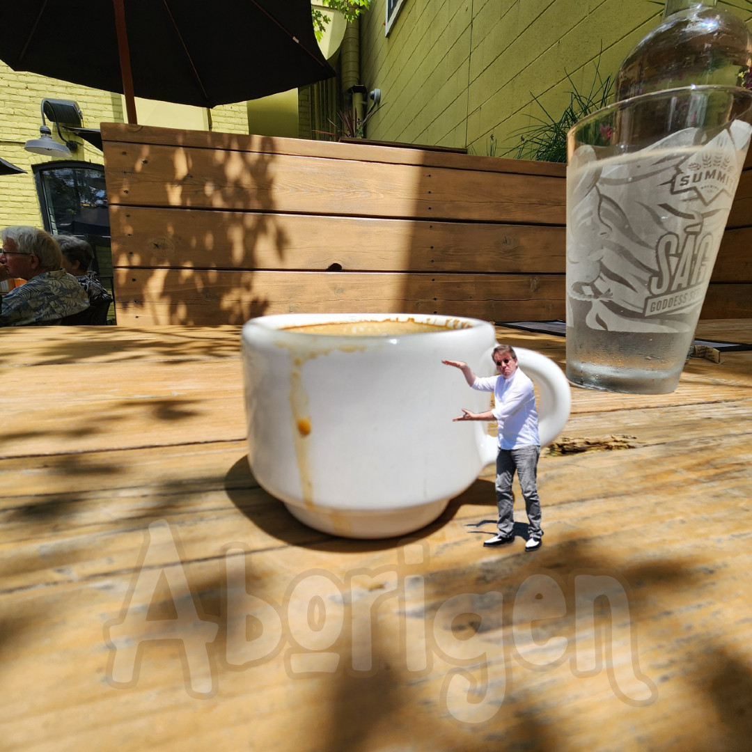 Tiny Aborigen stands by a demitasse of espresso, on a cafe table, gesturing for a cup even smaller than that.