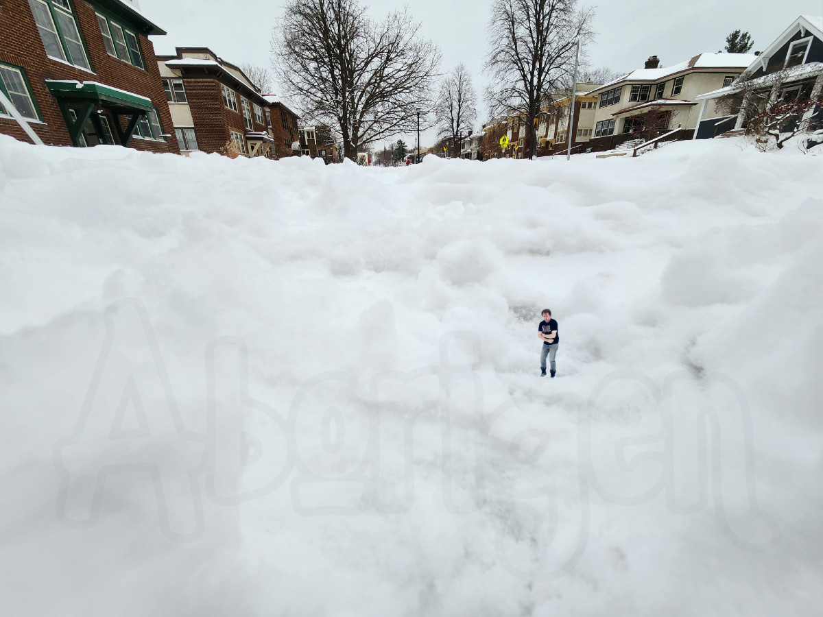 Low-angle shot of a snowy neighborhood, close-up shot of the footsteps along an unshoveled sidewalk. In one huge footstep, tiny Aborigen crouches, clutching his body for warmth.