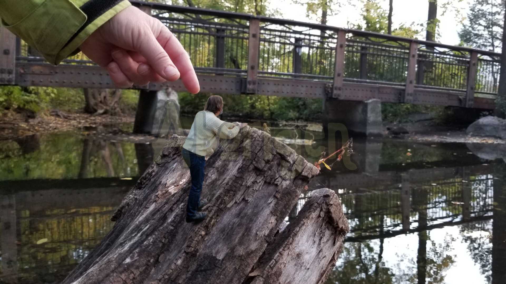 Tiny Aborigen is climbing a stump to look at a placid creek. A woman's gigantic hand reaches down to pluck him up.