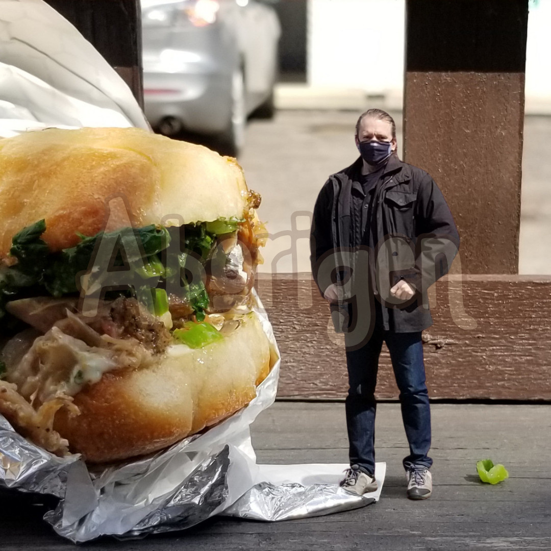 Tiny Aborigen stands beside a huge, delicious pork sandwich.