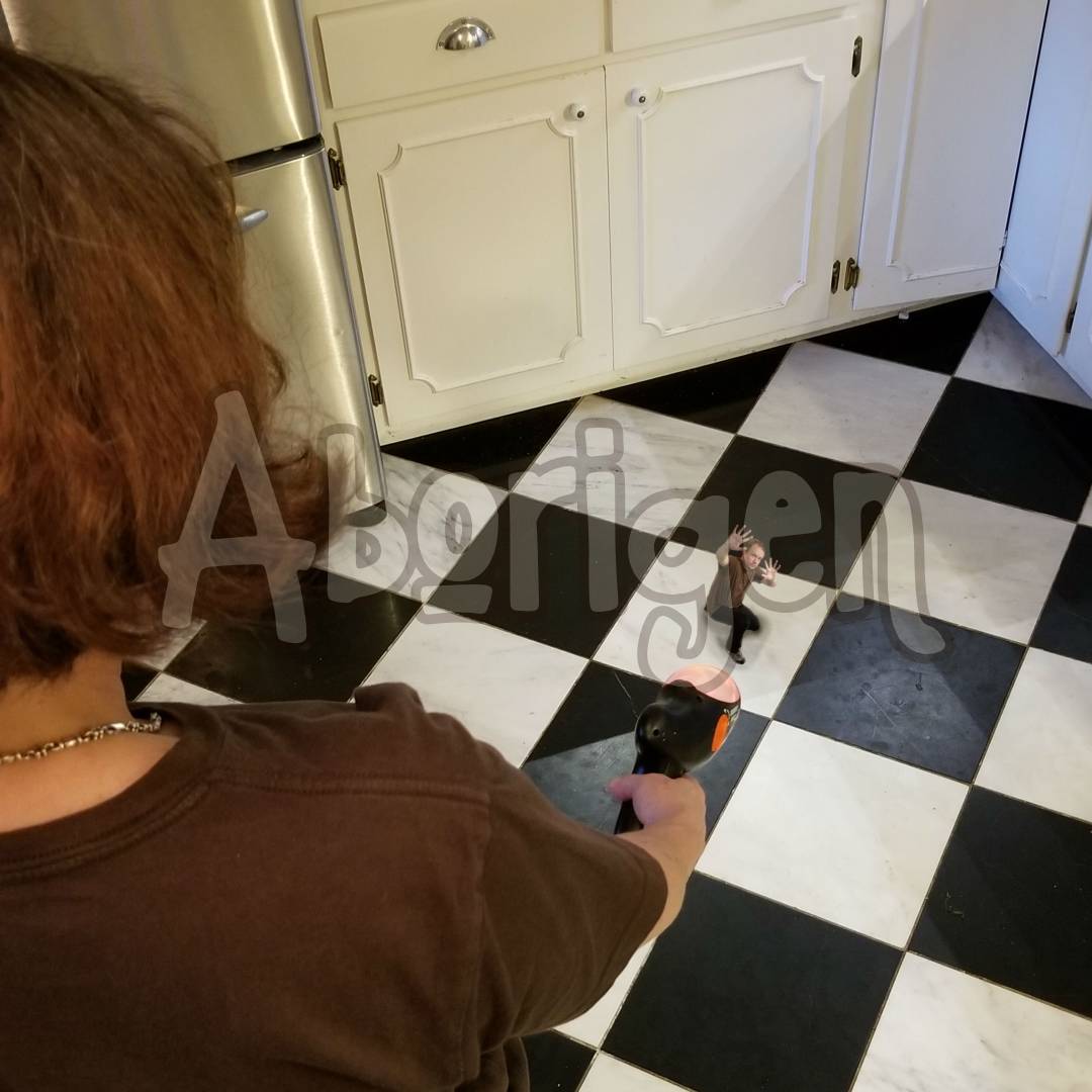 The camera looks down over the shoulder of a woman holding a shrink ray, pointing it at a shrunken man on a kitchen floor.